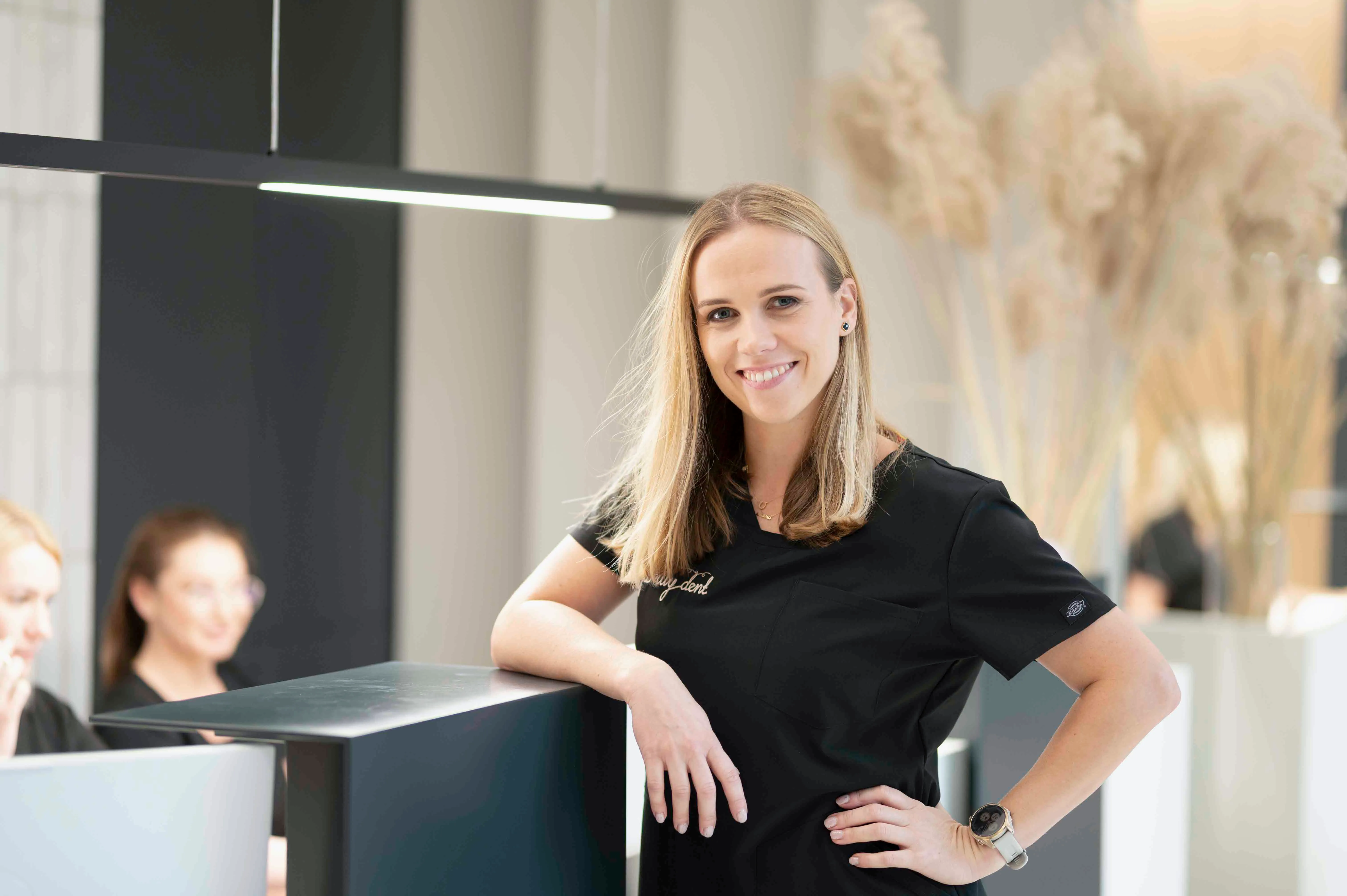 Uśmiechnięta pracowniczka kliniki stomatologicznej w eleganckim, minimalistycznym wnętrzu przy recepcji.